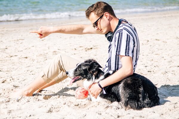 Mann am Strand zeigt in die Ferne, Hund sitzt daneben und schaut in die gleiche Richtung